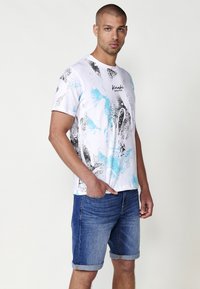 Bílý tričko s modro-černým abstraktním vzorem a logem "Kashii Brand". V kombinaci s ohrnutými džínovými šortkami.