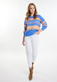 Mėlynas ir oranžinis raštuotas megztinis su laisvomis rankovėmis, derinamas su šviesiais džinsais ir rudos spalvos siaurais batais. Minkšta, tekstūruota megztos medžiagos struktūra.