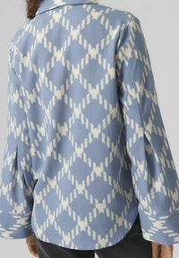 Blå mönstrad skjorta med vita diamantformer, gjord av texturerat tyg, med krage och vida ärmar. Bakvy visas.
