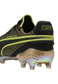 Scarpa da calcio nera e gialla con motivo scacchiera, suola traslucida iridescente e marchio distintivo sul tallone.