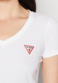 Camiseta blanca de cuello en V con un logo triangular rojo en el pecho que dice "GUESS" y un signo de interrogación. Textura de tela de algodón suave.