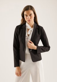 Schwarzer taillierter Blazer aus glattem Stoff mit Reverskragen, einer Knopfverschluss und dezenten Taschen, kombiniert mit einer weißen Bluse.
