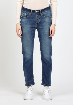 Person trägt ausgewaschene blaue Jeans mit geradem Bein, weißes Langarmshirt und weiße Nike-Sneaker, steht vor einem einfarbigen Hintergrund.