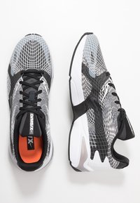 Scarpe da corsa bianco e nero con tomaia in rete che presenta un motivo a griglia, interno arancione, lacci neri e una suola bianca imbottita.