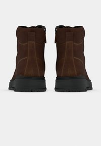 Stivali alla caviglia in suede marrone con suola in gomma nera, dotati di una cerniera laterale e dettagli di cucitura visibili sulla parte superiore.