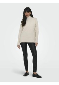 Pull à col roulé crème avec des poignets et un ourlet côtelés, associé à un pantalon noir ajusté et des mocassins noirs, présentés sur un fond clair.