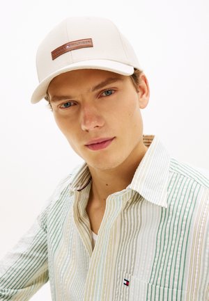 Ung mand med blå øjne iført en beige Tommy Hilfiger-kasket og en flerstribet skjorte med knapper og et lille logo på lommen.