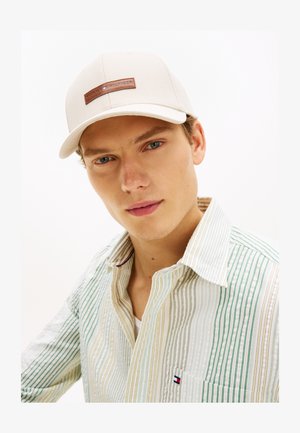 Jeune homme aux yeux bleus portant une casquette beige Tommy Hilfiger et une chemise à boutons à rayures multicolores avec un petit logo sur la poche.