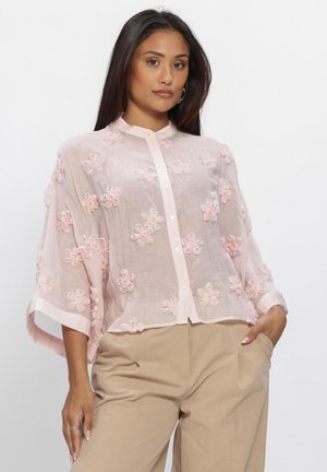 Vrouw die een losse lichtroze blouse met knopen en bloemrijke borduurwerk draagt, gecombineerd met beige broek, poserend met één hand in de zak.