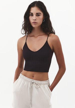 Mujer joven con cabello rizado y oscuro que lleva una camiseta negra corta y pantalones beige claro con cordón, sobre un fondo blanco.