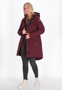 Bordeauxkleurige parka met capuchon, voorzien van een voorrits en drukknoopsluitingen, zijzakken en een met grijs gevoerde capuchon; gedragen over zwarte jeans en lichte schoenen.