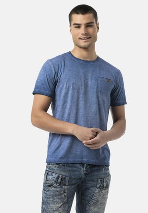 Young man with short hair wearing a blue t-shirt and faded cargo jeans, standing with hands clasped in front, smiling gently.