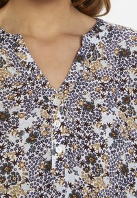 Chemisier à fleurs boutonné avec un décolleté en V, tissu blanc orné de motifs floraux bleus, bruns et jaunes, et trois boutons décoratifs.