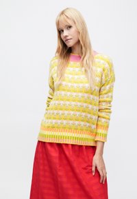 Pletený sveter v žltej farbe s ružovými akcentmi, zdobený kvetinovým vzorom a horizontálnymi pruhmi. Kombinovaný s voľnou červenou sukňou.