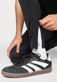 Μαύρα και λευκά παπούτσια Adidas Predator με καπακι από σουέντ στη μύτη, καουτσούκ σόλα και κορδόνια. Φοριούνται με μαύρο στενό παντελόνι και λευκές κάλτσες.