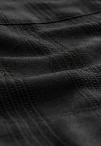 Sort stof med en subtil hævet gittermønster og bløde folder, der fremhæver tekstur og vævningsdetaljer.