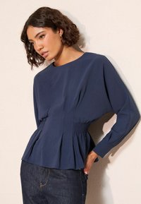 Tumesinine, pika varrukaga topp, millel on volditud, smock-kere, ümmargune kaelus ja tekstuuriga kangas, kombineeritud tumedate teksadega.