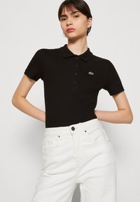 Polo shirt nero a coste con colletto e bottoni, caratterizzato da un logo ricamato verde. Abbinato a pantaloni in denim bianchi a vita alta.