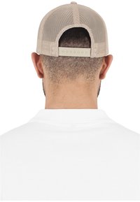Cappellino da baseball in rete beige con cinturino regolabile sul retro, posizionato su uno sfondo di camicia bianca. La texture appare traspirante.