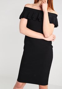 Czarna sukienka bez ramiączek z falbaną przy dekolcie. Dopasowany krój z długością do kolan, wykonana z gładkiego, elastycznego materiału.