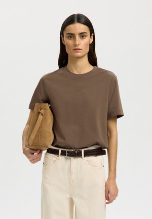 Femeie cu părul lung și închis la culoare, purtând un tricou maro și pantaloni bej, ținând o geantă de mână din piele întoarsă de culoare cămilă sub braț, pe un fundal simplu.