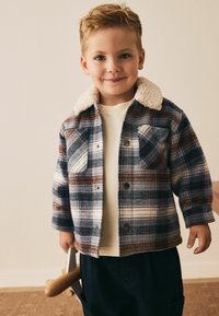 Bordas jaku ar zilu, sarkanu un baltu rūtainu rakstu, ar krēma krāsas mākslīgā kažokādas apkakli un divām krūšu kabatām, valkāta virs gaismas džempera.