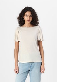 Šviesiai ruda trumparankovė marškinė su susirinkusiomis pūstomis rankovėmis ir plačia apykakle, pagaminti iš minkštos, lengvos medžiagos. Dėvimi su mėlynomis džinsinėmis kelnėmis.