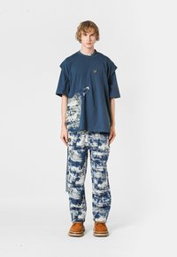 Joven de pie, vistiendo una camiseta azul de gran tamaño con un estampado abstracto, pantalones a juego azul y blanco con patrones, y zapatos marrones.