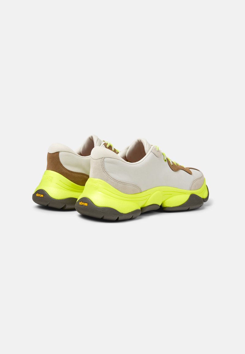Baskets sportives avec une tige synthétique beige, des accents en suède marron, une semelle jaune vif et des lacets verts, dotées d'une semelle extérieure en caoutchouc texturé.