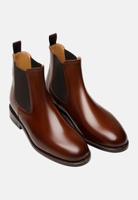 Bottines chelsea en cuir marron avec des panneaux latéraux élastiques, finition lisse et brillante, et un talon plat. Équipées de languettes pour un enfilage facile.