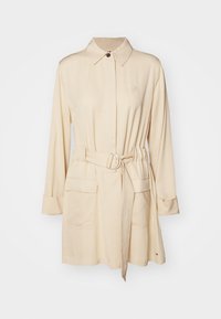 Robe chemise beige ceinturée avec un col, des manches longues, deux poches avant et un détail logo sur la poitrine. Tissu doux et coupe ajustée.