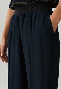 Marineblauwe wijde broek van een gestructureerde stof, met een zwarte elastieken tailleband en zijzakken, getoond van de taille tot beneden.