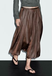 Vrouw in een glanzende bruine midi-jurk, grijze lange mouw top en zwarte platte sandalen die over een zwarte vloer loopt tegen een witte muur.