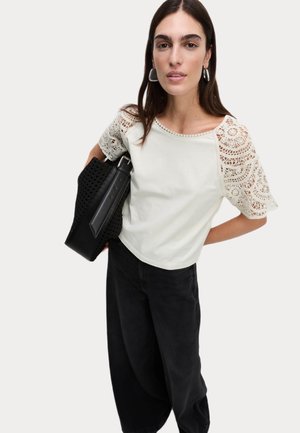 Top bianco cropped con maniche in pizzo, abbinato a pantaloni oversize neri e una borsa a tracolla nera con texture. Orecchini a cerchio in argento accentuano il look.