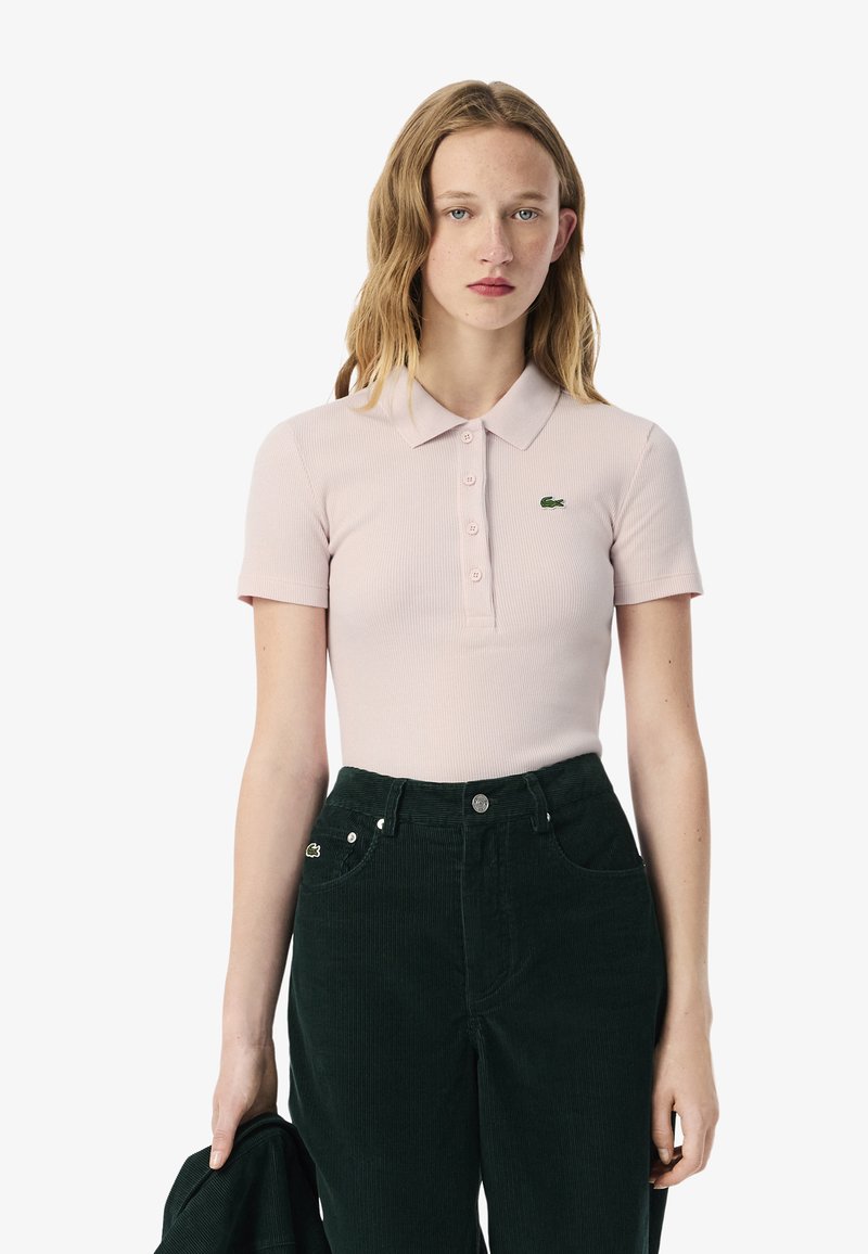 Vaaleanpunaine röyditetty polo-paita lyhyillä hihoilla ja nappilistan kanssa, jossa on pieni vihreä Lacosten logo. Yhdistetty tummanvihreisiin samettihousuihin.