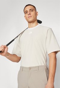 Cămașă de golf crem cu guler, cu mâneci scurte, având trei dungi albe pe piept. Asortată cu pantaloni de golf bej. Ținând un club de golf.