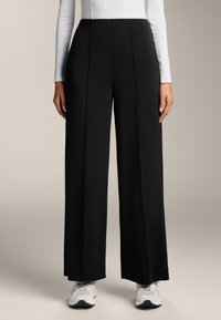 Fekete, bő szárú nadrág sima anyagból, elöl redőzött és magas derekú, fehér sportcipővel párosítva.