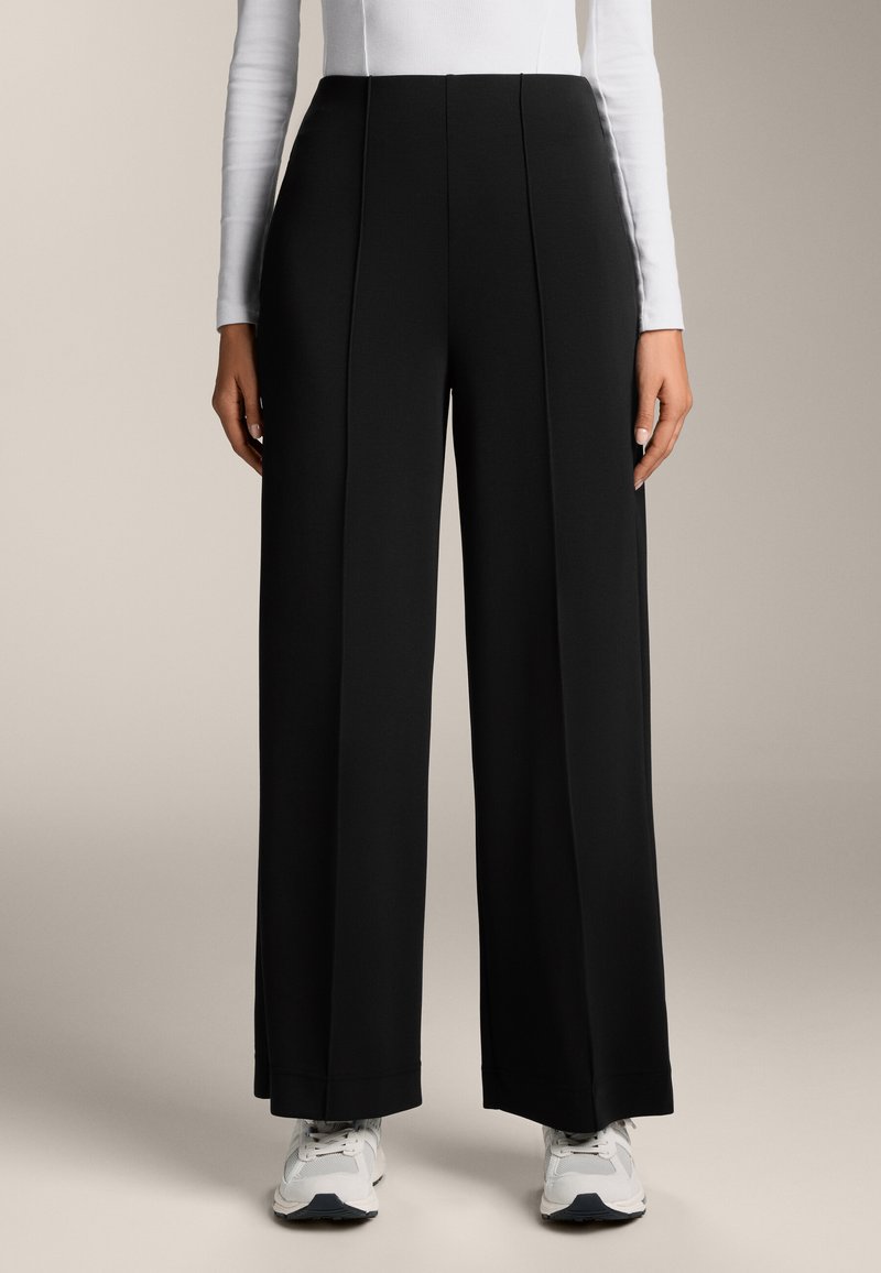 Fekete, bő szárú nadrág sima anyagból, elöl redőzött és magas derekú, fehér sportcipővel párosítva.