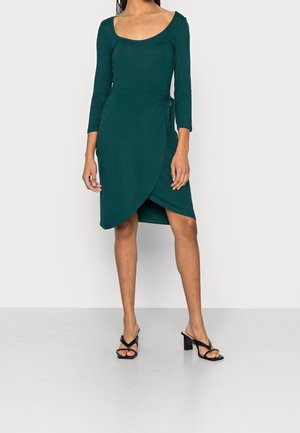 Teal wrap dress with three-quarter sleeves, scoop neckline, and side tie detail. Paired with black heeled sandals. Smooth texture.