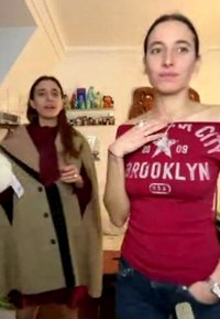 Deux femmes à l’intérieur ; l’une ajuste un collier en forme d’étoile sur un top Brooklyn rouge, l’autre porte un manteau beige et tient une étiquette dans une pièce avec des étagères remplies de figurines.