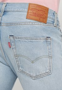 Jeans de mezclilla azul claro con un parche de cuero con marca grabada, costuras distintivas en los bolsillos traseros y una pequeña etiqueta de logo roja.