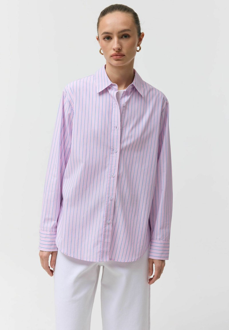 Kobieta ubrana w jasnofioletową koszulę na guziki z cienkimi niebieskimi pionowymi paskami oraz białe spodnie, stojąca na tle jednolitego białego tła.