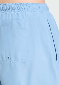 Světle modré šortky s elastickým pasem, bočními sklady a zadními náprsními kapsami s detailem knoflíkové dírky. Hladká textura materiálu.