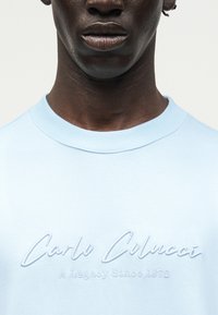 Κοντινό πλάνο ενός ατόμου που φοράει ένα ανοιχτό μπλε φούτερ με κεντημένα τα λόγια "Carlo Colucci" και "A Legacy Since 1978" στο στήθος.