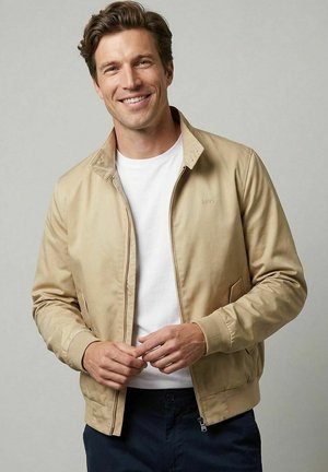 Lächelnder Mann, der eine beige Jacke über einem weißen Hemd und dunklen Hosen vor grauem Hintergrund trägt, die Hände locker an der Taille verschränkt.