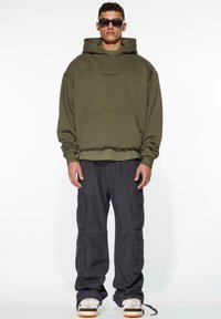 Olivengrønn oversized hettegenser med kengurulomme og brodert logo, parret med svarte cargo-bukser som har flere lommer og snøring.