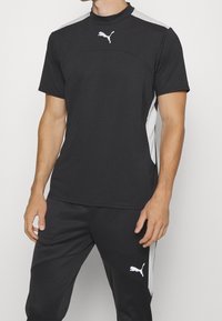 Top deportivo negro con un cuello alto y mangas cortas. Presenta acentos grises a lo largo de los hombros y un logo blanco de Puma en el pecho.