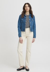 Mėlyna džinsinė striukė su sagomis priekyje, dviem krūtinės kišenėmis ir trumpu kirpimu. Dėvima ant juodai baltai dryžuotos marškinėlių ir smėlinio spalvos džinsų.