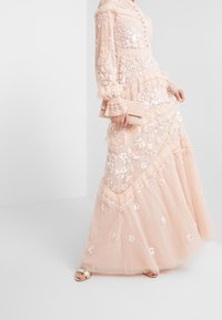 Lång, ljusrosa klänning i genomskinligt tyg med blomstermodel och paljetter. Har volangärmar och ett lager på lager-skjort.