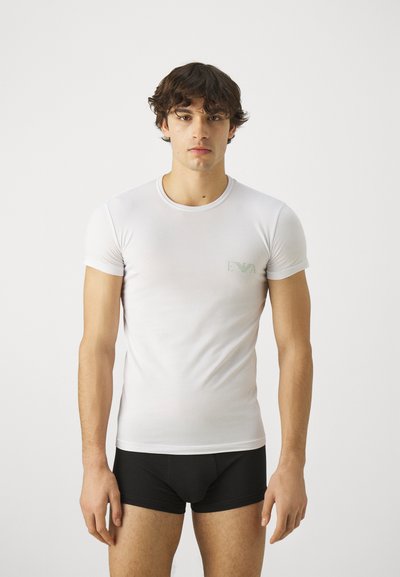 Camiseta blanca de manga corta con un diseño ajustado y un logo verde en el pecho. El modelo lleva calzoncillos bóxer negros y está de pie frente a un fondo neutro.